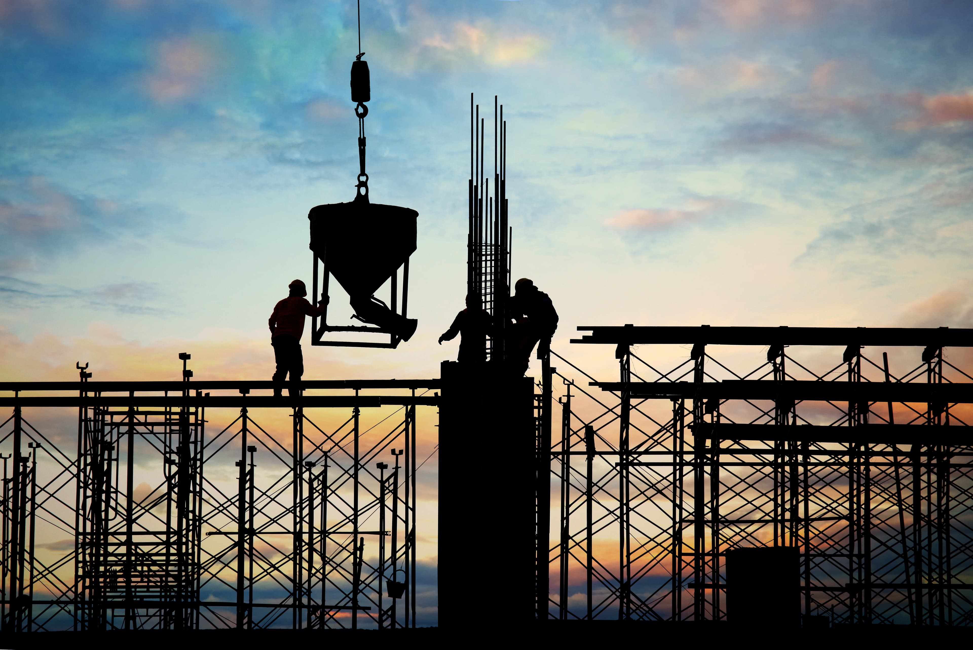 Workers on a scaffolded building under construction at sunset, silhouetted against a concrete bucket lifted by a crane